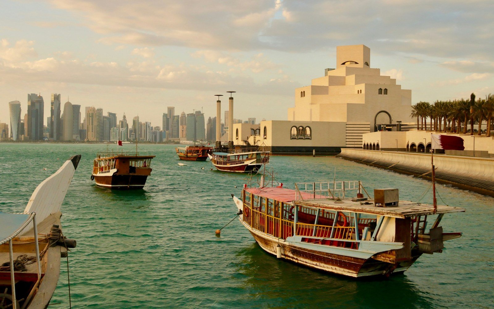 Wooden dhow boats moored on green water in front of a low sand-coloured museum building, with a line of tall towers on the far shore under a hazy sky