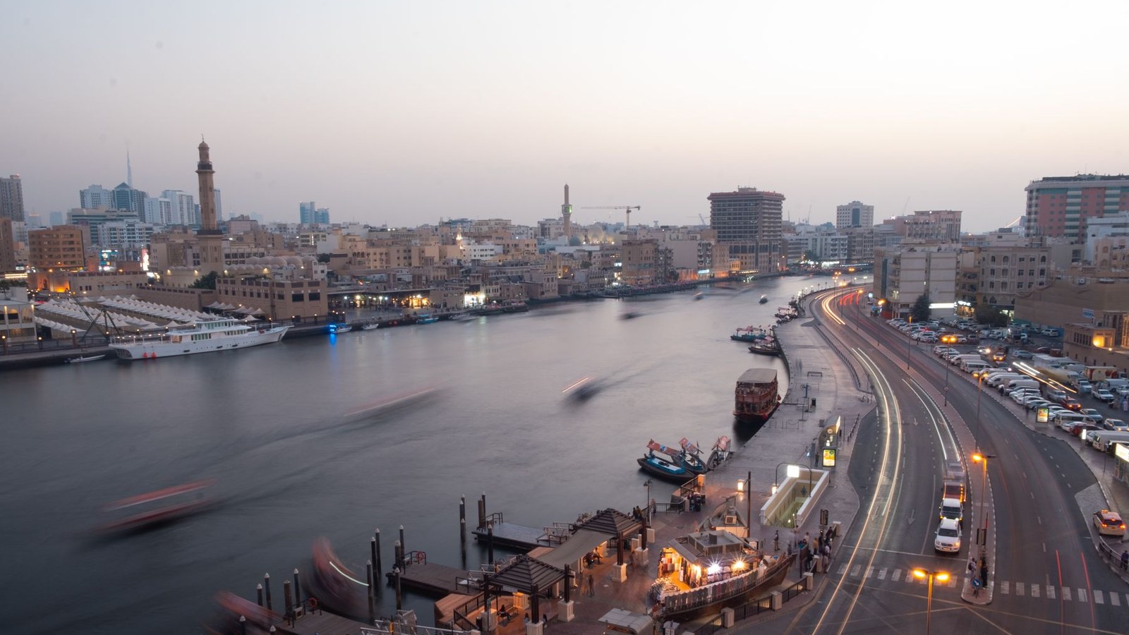 Dubai Creek at dusk, with lit waterfront buildings, moored abras and traffic on the road beside the water