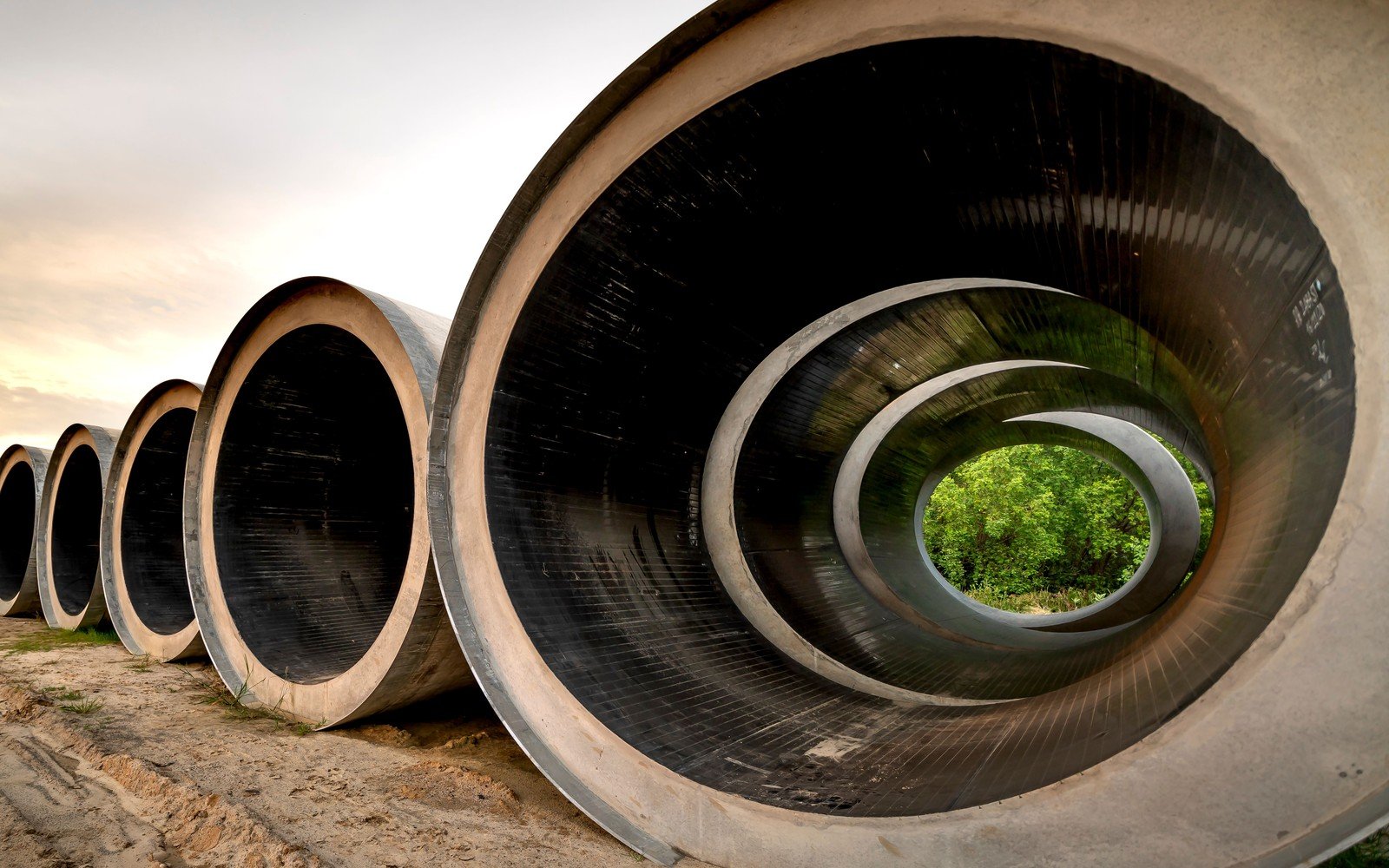 A line of very large precast concrete pipes lying end to end on bare ground at dusk, the nearest one open to the camera so the bore runs away through the pipes behind it