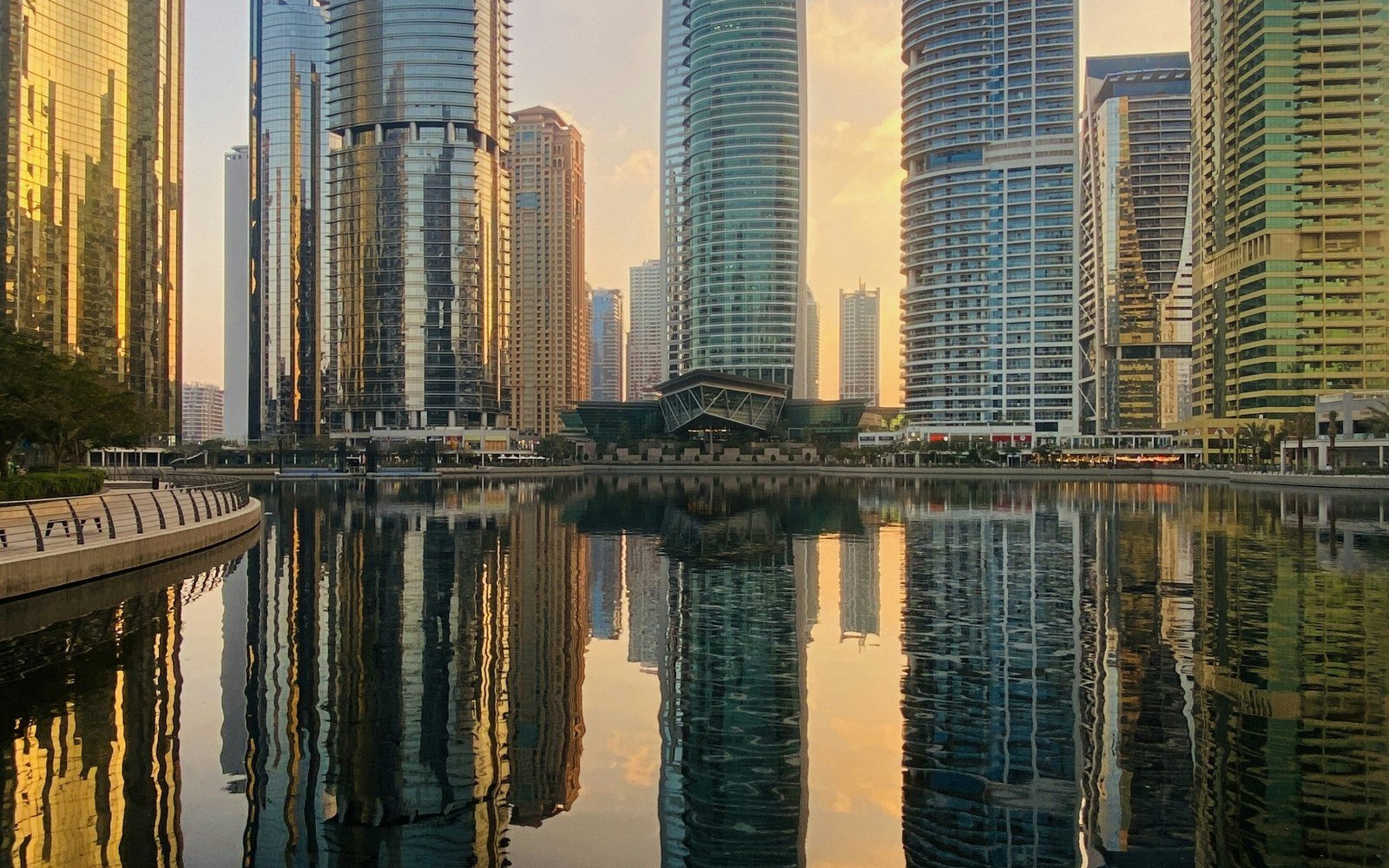 A cluster of glass-clad residential and office towers around a still lake at first light, with a low pavilion on the far bank and the whole group reflected in the water