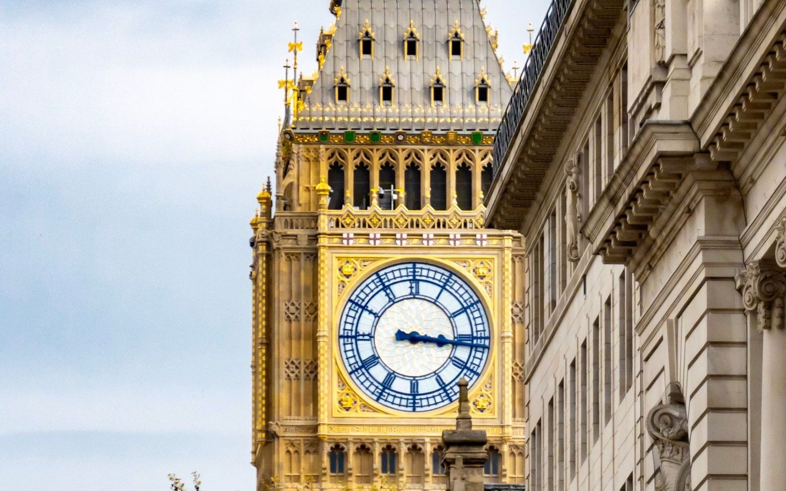 The clock face and gilded upper stage of the Elizabeth Tower in daylight, framed against the stone flank of a neighbouring building