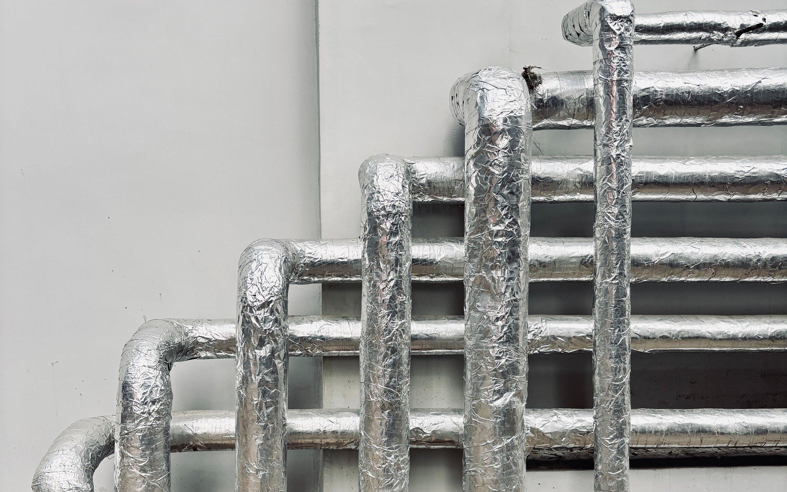 Insulated pipework clad in bright foil running up and across a plain pale wall, three vertical risers turning through swept bends into a stack of horizontal runs behind a plain louvred panel