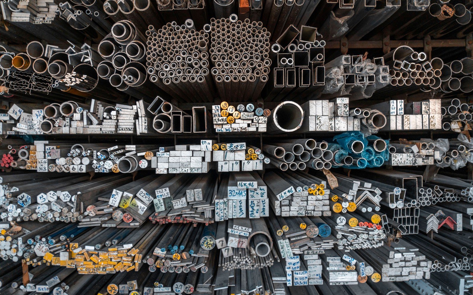 Racked metal stock seen end on, round tube, square and rectangular hollow section, flat bar and angle sorted by size across several shelves, each bundle carrying a small handwritten size label