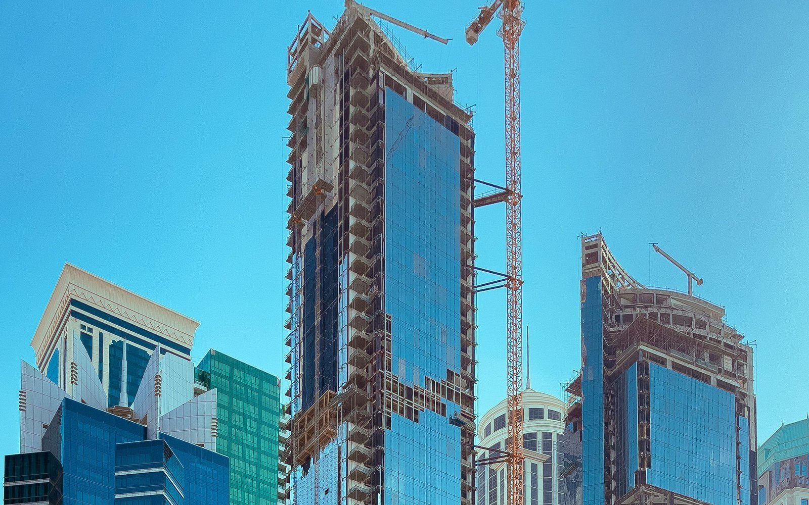 Two high rise towers under construction with a tower crane between them in the West Bay district of Doha, with completed glass and stone towers to the left under a clear sky