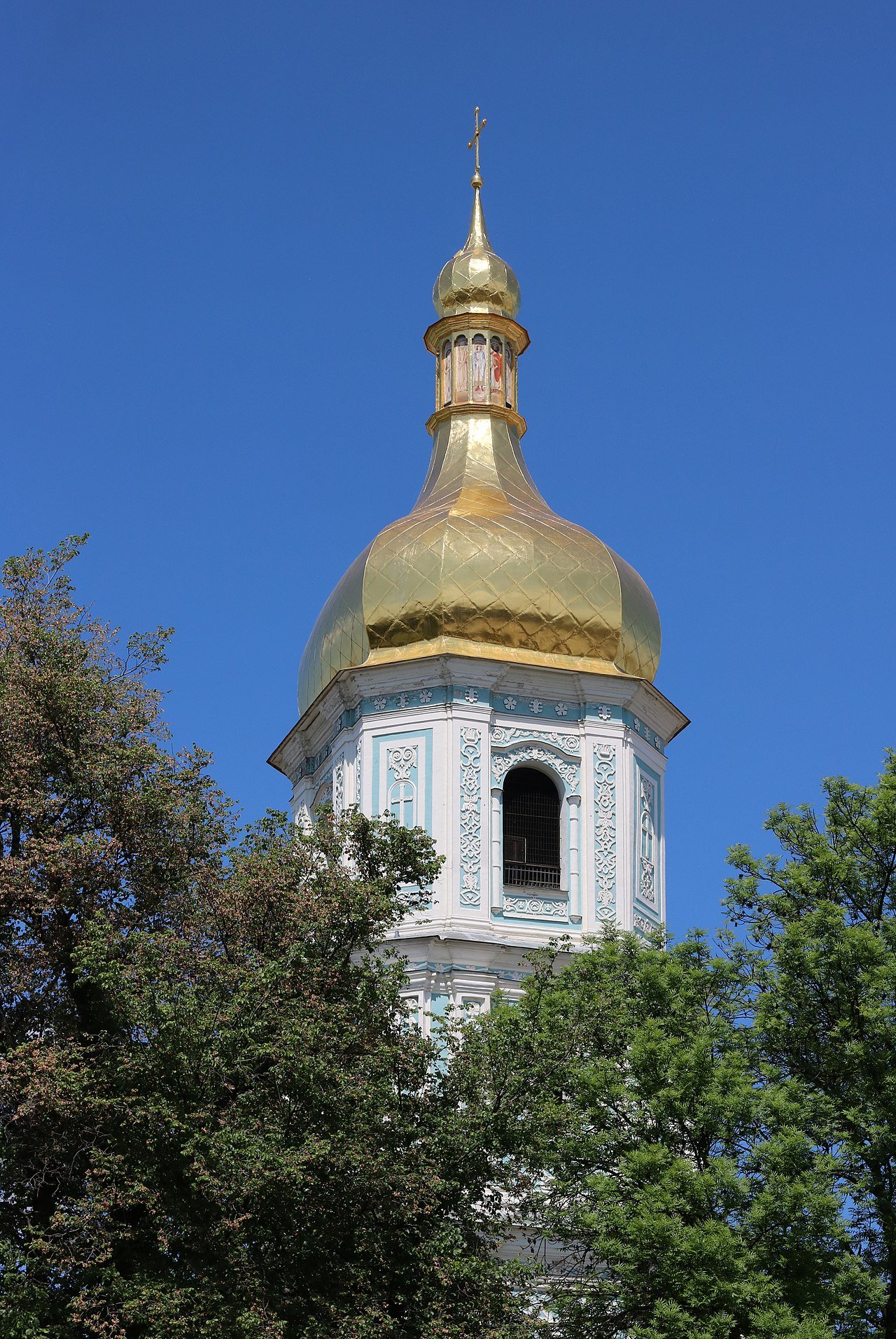 A gilded onion dome and white belfry rising above summer trees against a clear sky