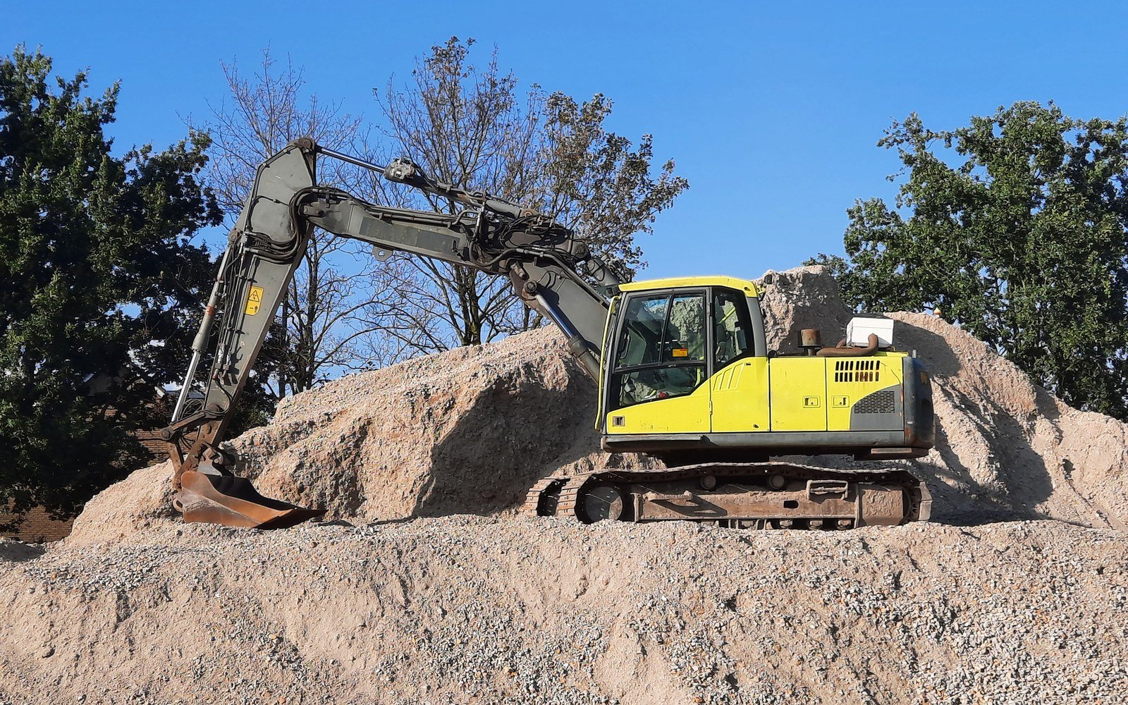 A yellow tracked excavator standing on a mound of crushed stone with its boom raised and the cab empty, bare trees and clear sky behind