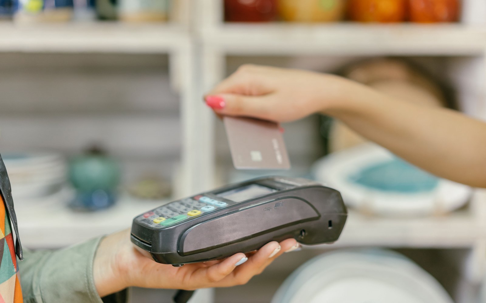 A hand holding an unmarked bank card above a black handheld card reader held out by a shop assistant