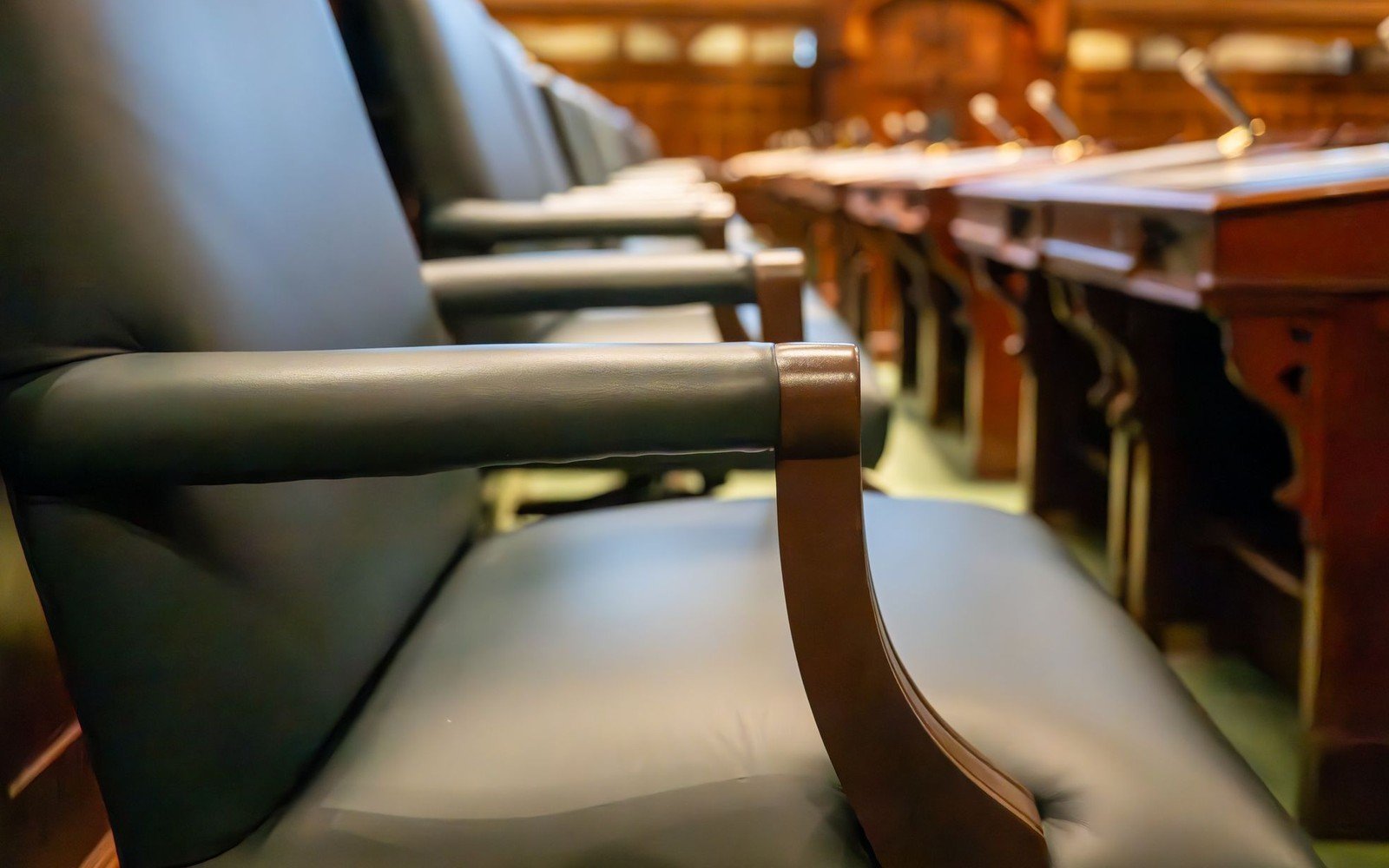A row of empty leather chairs along a timber courtroom desk, small microphones on stands running away down the bench and the far end of the room out of focus