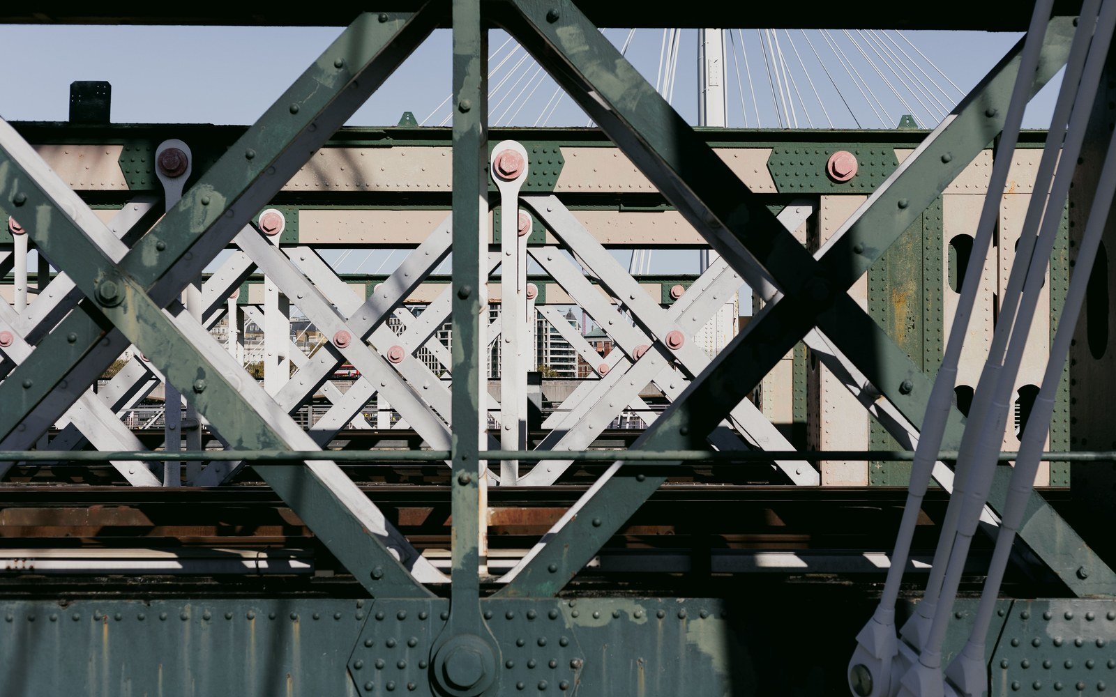 The riveted steel lattice of a railway bridge photographed from the deck, cross bracing and gusset plates in the foreground and city buildings beyond