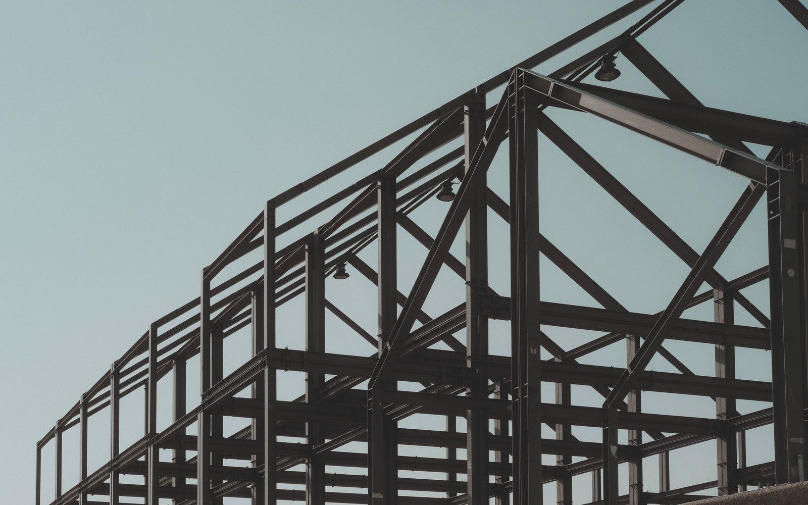Structural steel frame of a large single storey building seen from below against a pale sky, bolted portal frames and diagonal bracing carrying a row of downlights, with no cladding yet fixed