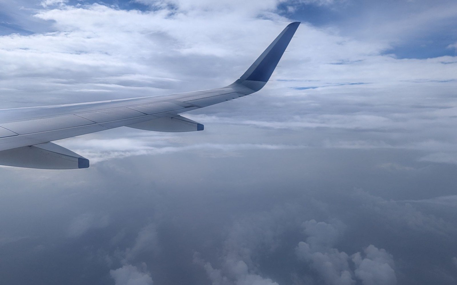 The wing of an airliner seen from a cabin window above a layer of cloud, with a second cloud deck below and pale sky at the horizon