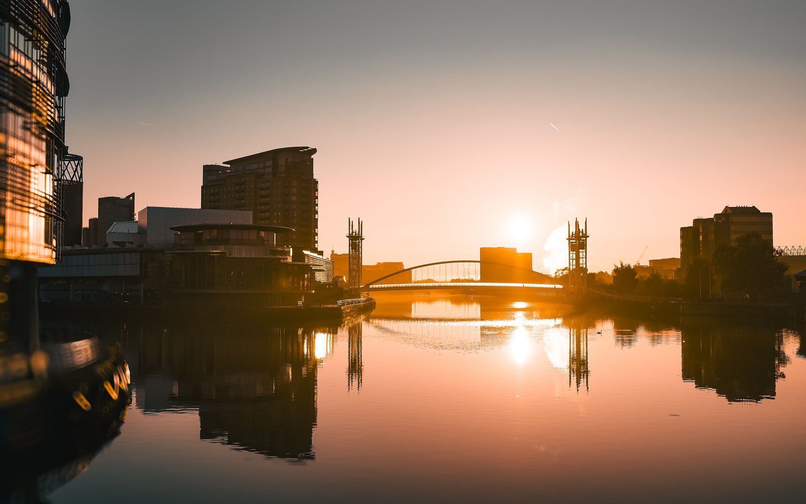 Sunrise over the water at Salford Quays, with a footbridge arch and a lift bridge tower in silhouette against an orange sky