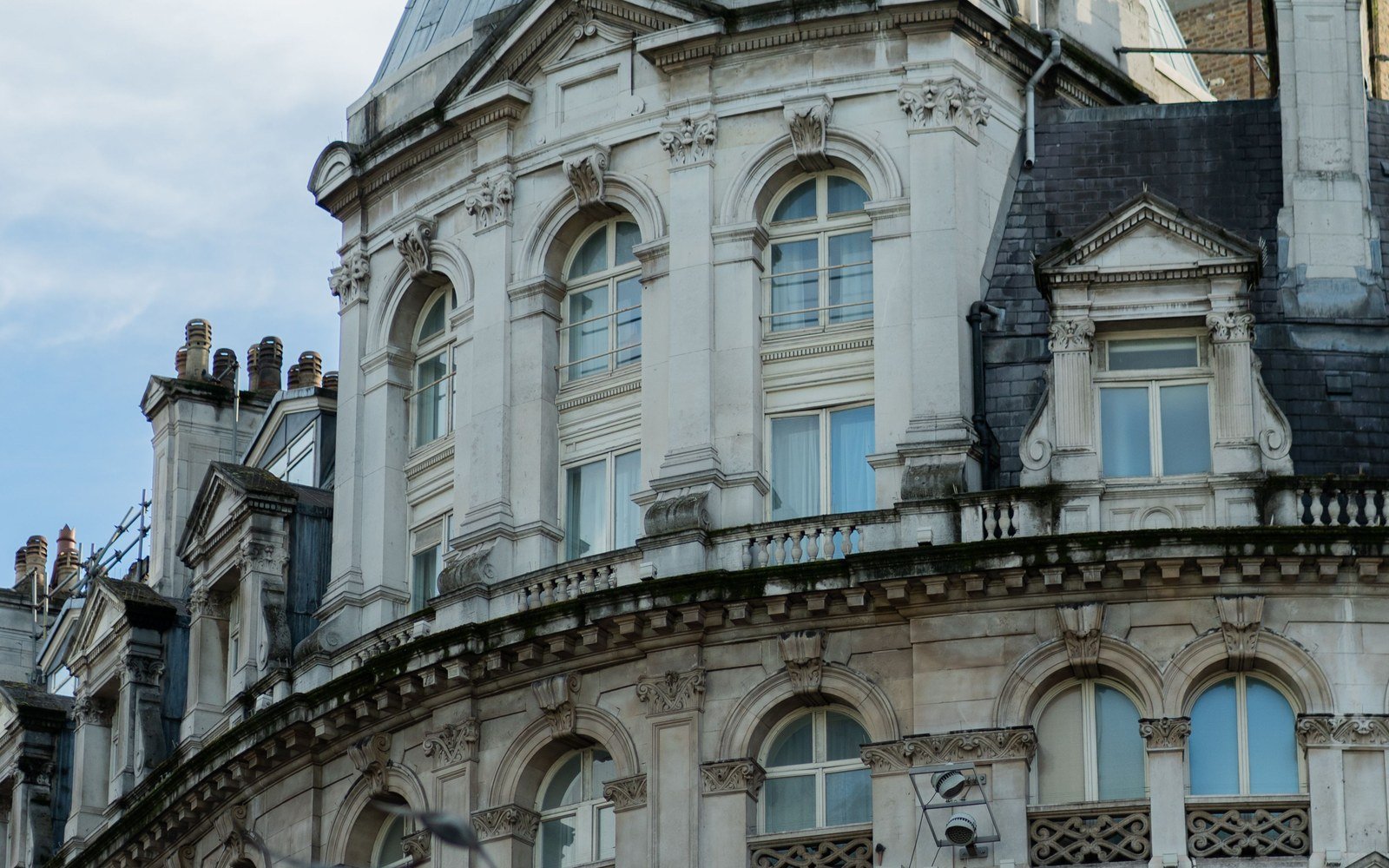 The upper storeys of a Victorian stone office building in London, with a domed corner turret, sash windows and a slate mansard roof against pale sky