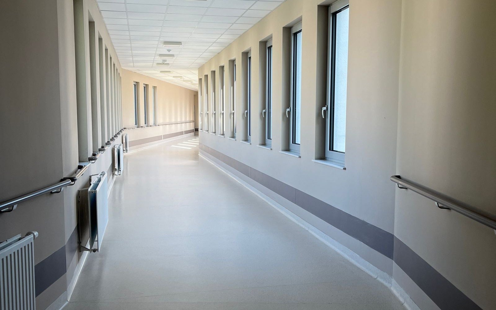 An empty hospital corridor with a run of tall windows down one side, handrails at waist height on both walls and a pale resin floor