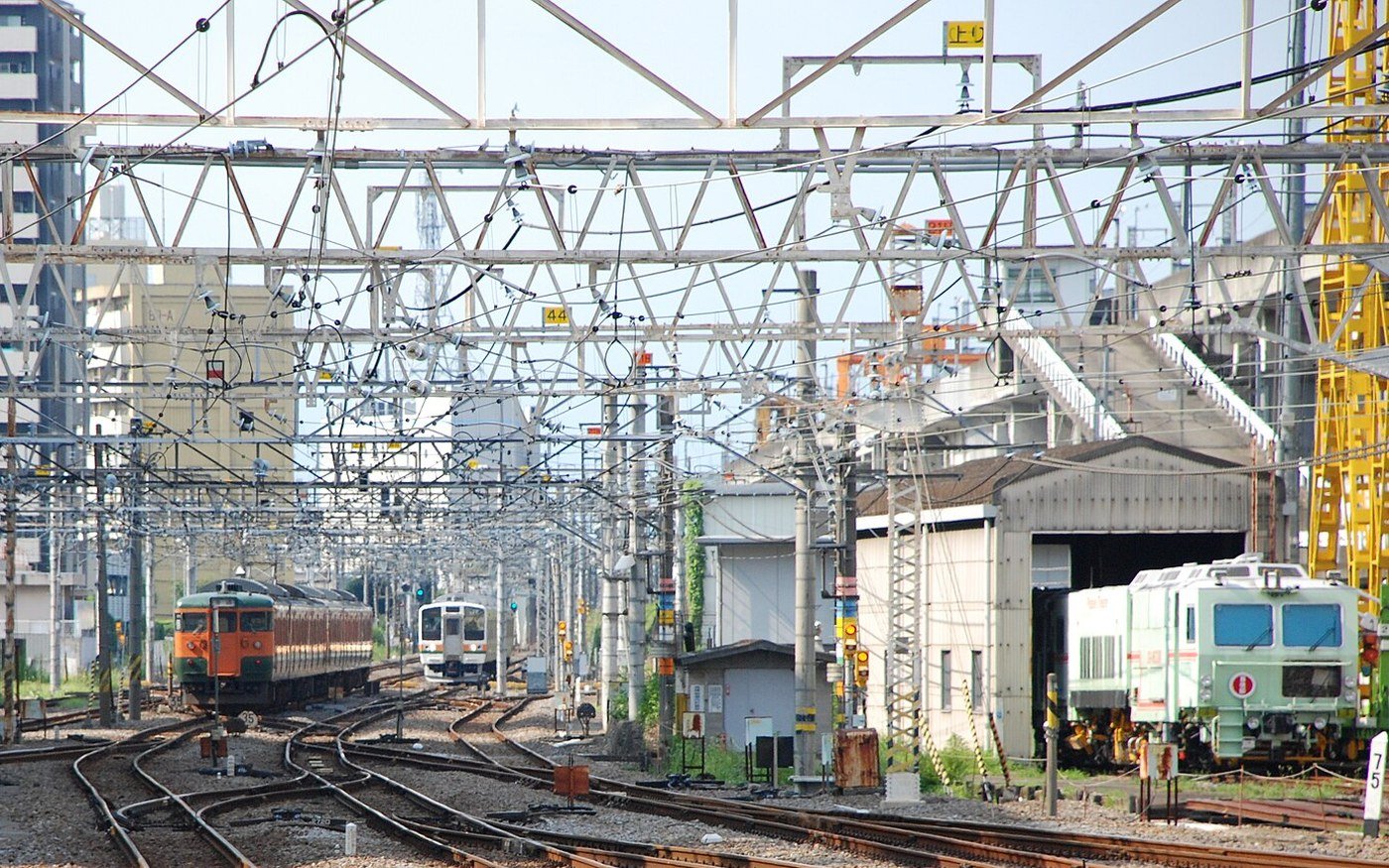 Several railway tracks converging through a station throat in urban Japan, ballast and point work in the foreground, overhead line gantries above the running lines and two stationary trains standing in the distance