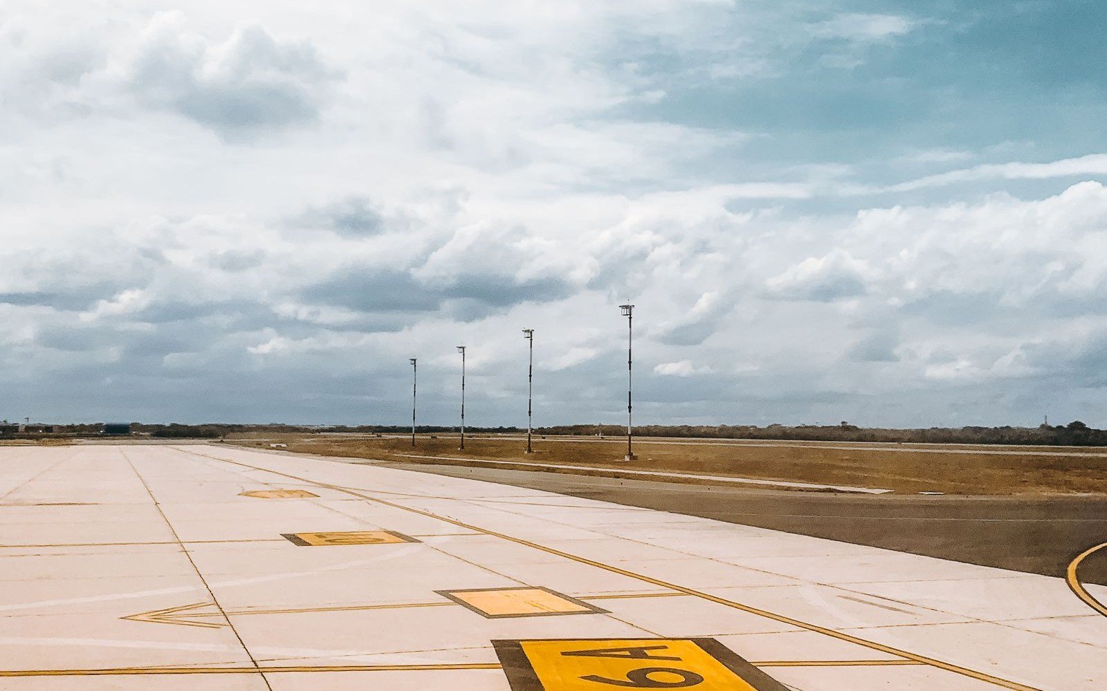 An empty runway with painted threshold markings running away towards approach lights and open ground under a cloudy sky