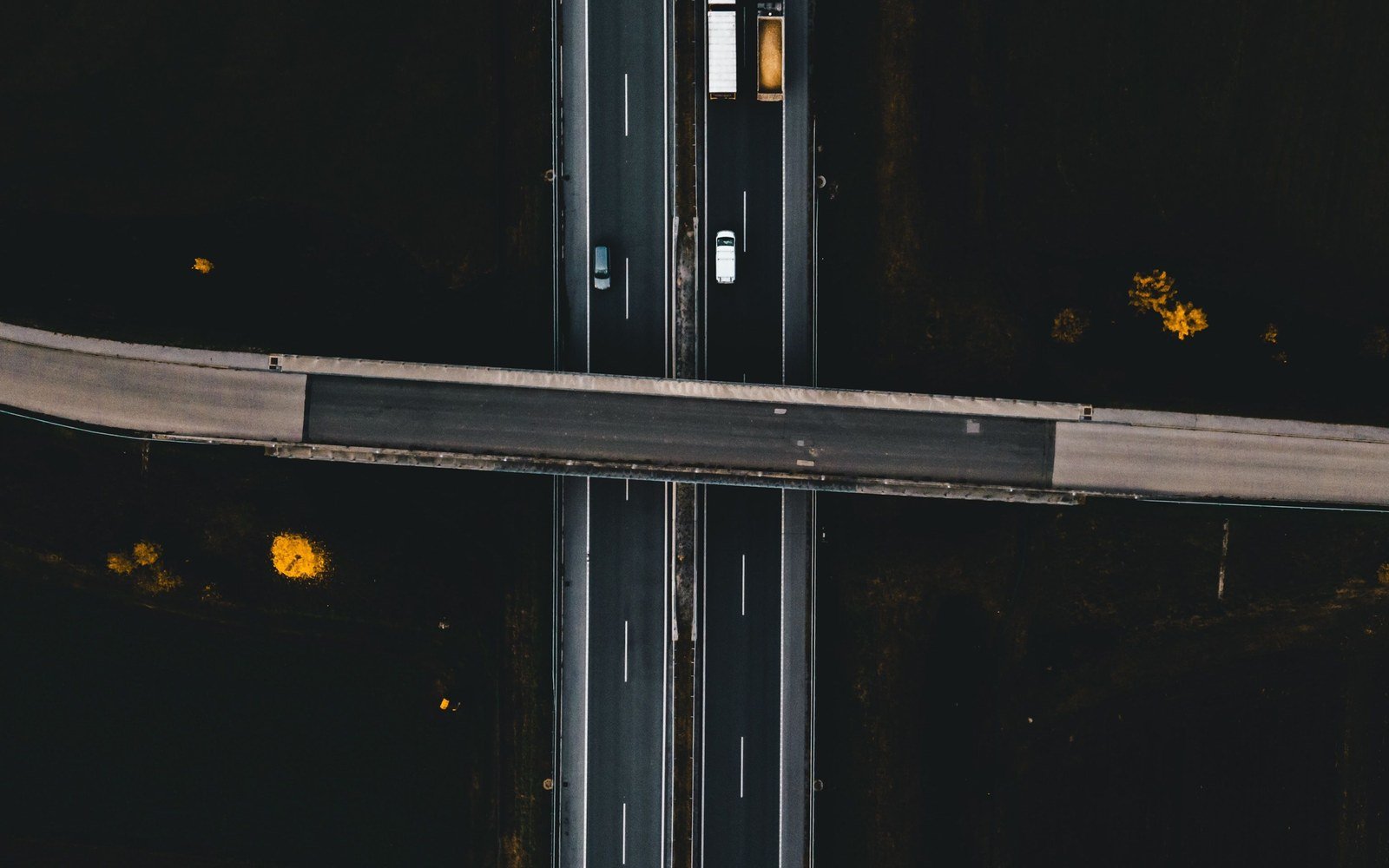 A stretch of dual carriageway photographed straight down from the air, two carriageways separated by a central reserve running under a plain concrete overbridge, a handful of vehicles spaced along the lanes and dark unlit ground either side