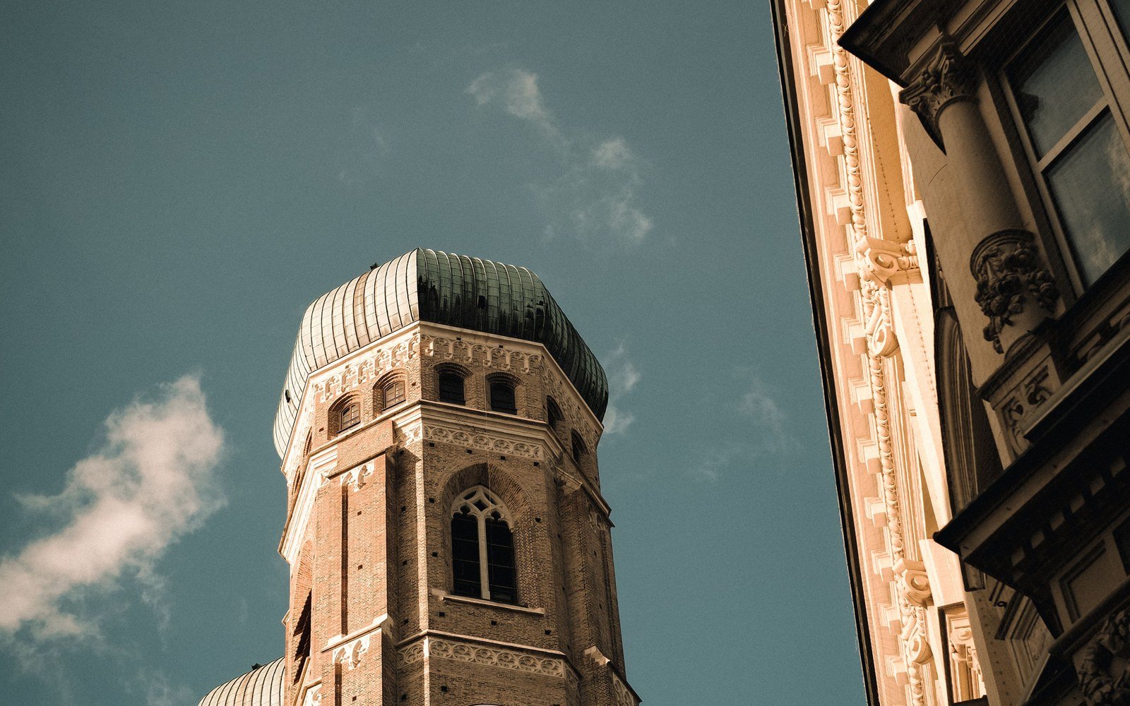 A low angle photograph of the brick tower and green copper dome of the Frauenkirche in Munich against a clear sky, with the stone facade of a neighbouring building at the right edge of the frame
