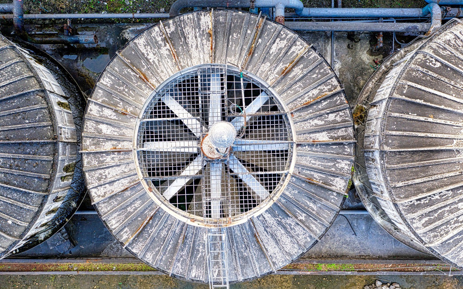 Overhead view of three large weathered metal ventilation fans set into an industrial roof, with pipework running across the frame