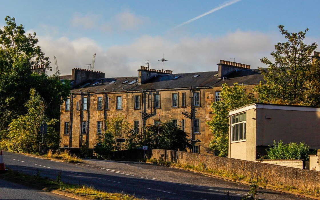 A terrace of sandstone tenements with slate roofs and chimney stacks beside a road, with construction cranes on the skyline behind