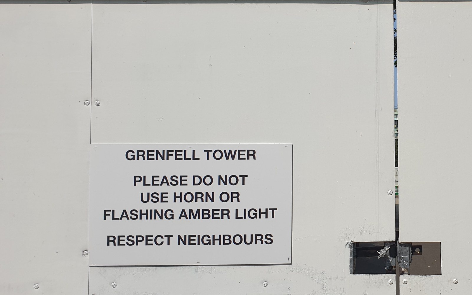 A white painted site hoarding carrying a printed notice headed Grenfell Tower, asking drivers not to use a horn or a flashing amber light and to respect neighbours
