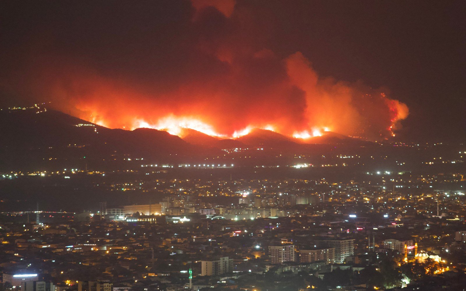 A line of fire burning along a dark ridge above the lights of a city at night