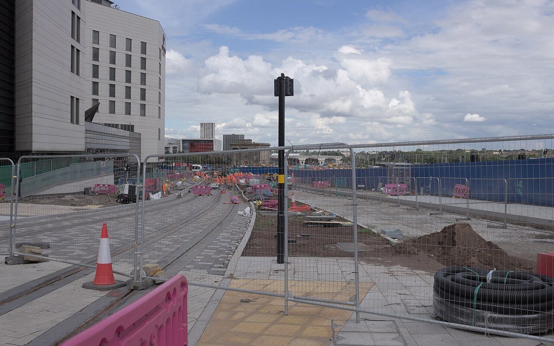 A city centre rail construction site behind temporary fencing, with track slab, ducting and a hoarded compound under a cloudy sky