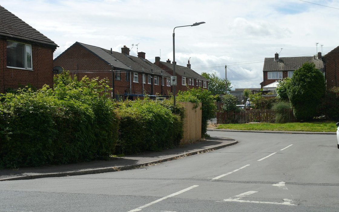 A row of brick local authority houses with tiled roofs, chimneys and hedges along a quiet residential road
