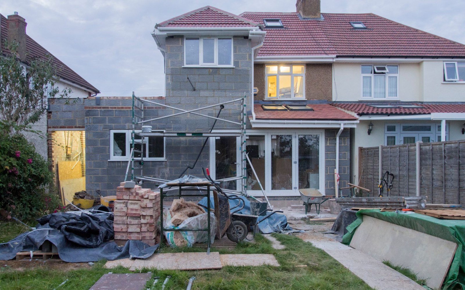 A rear extension under construction at a suburban house, with blockwork walls, scaffolding and materials stacked across the garden