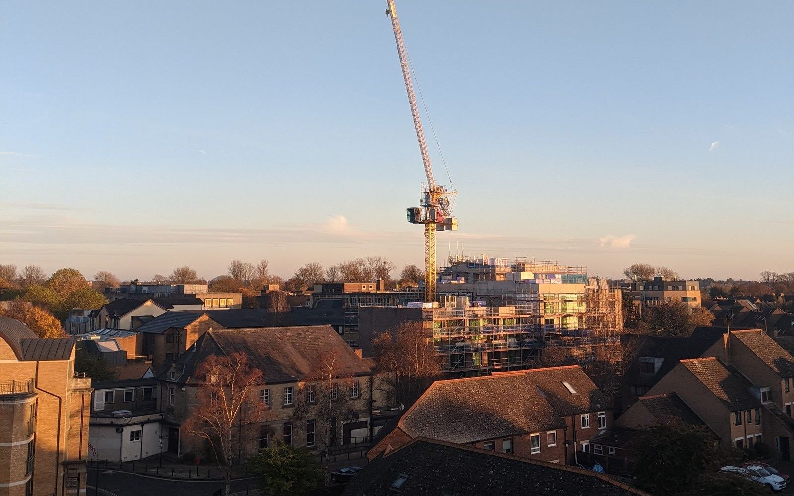 A tower crane standing over a scaffolded residential block among tiled roofs in an Oxfordshire town at sunset