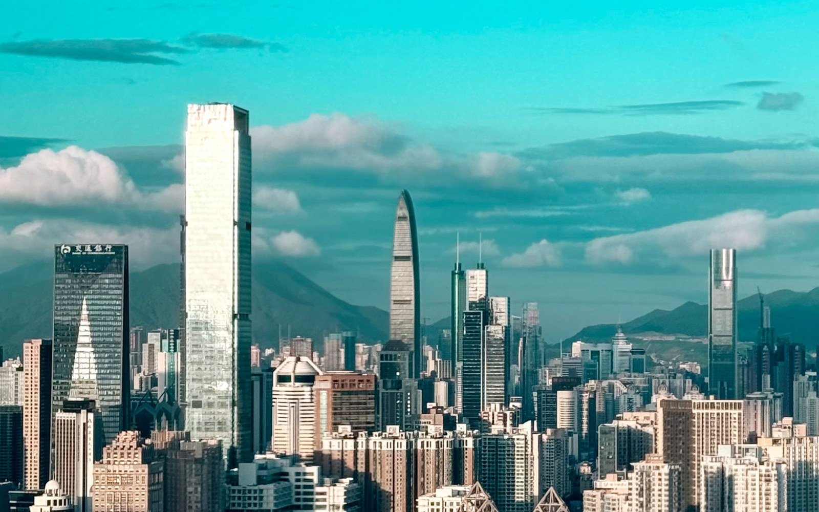 A daylight view of the Shenzhen skyline, a cluster of tall towers of different heights rising above a dense lower city with hills and cloud behind