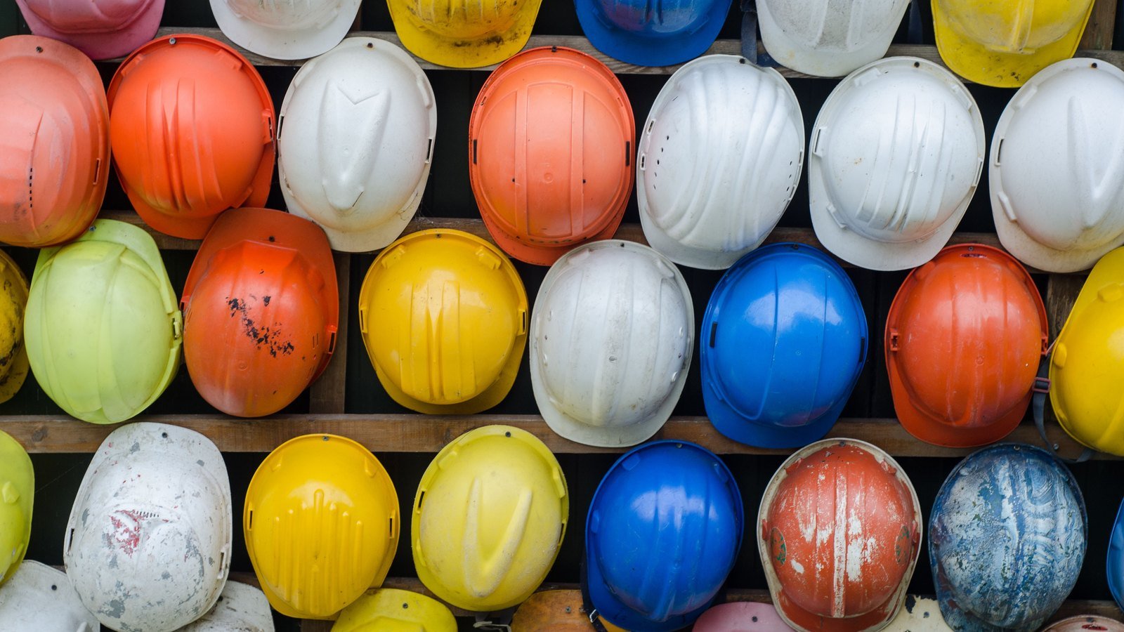 Rows of well used safety helmets in white, yellow, orange, blue and green stacked on wooden shelves