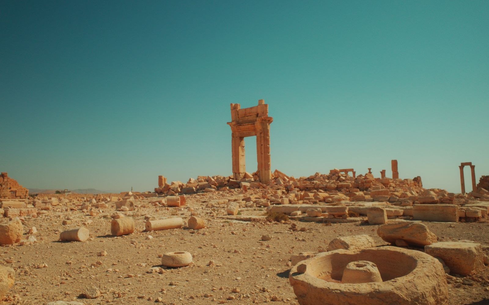 A standing stone gateway on open stony ground at Palmyra in the Syrian desert, with fallen column drums and cut blocks scattered across the foreground under a clear sky
