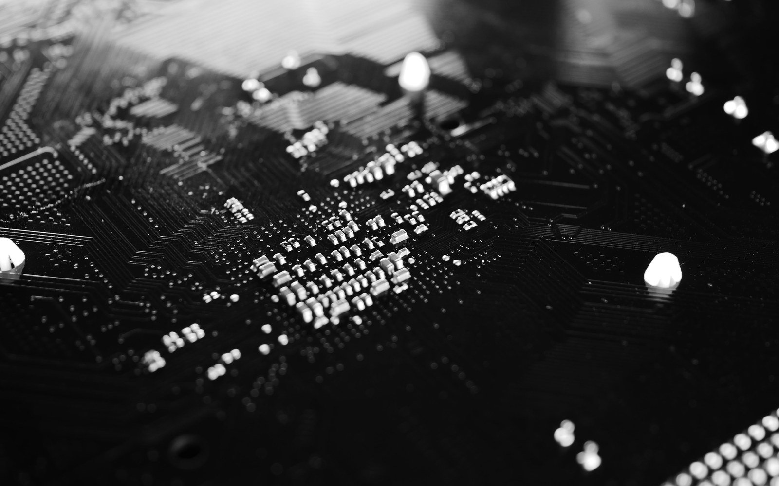 A black and white macro photograph of a bare printed circuit board at a shallow angle, rows of small surface mounted components catching the light and etched tracks running away into shadow