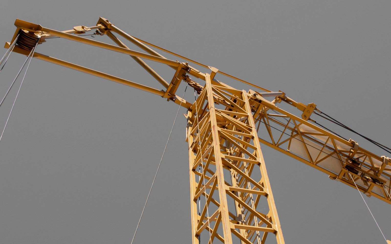 Upper section of a yellow lattice tower crane against a flat grey sky, the jib running across the frame with its pendant cables and hoist rope, seen from below