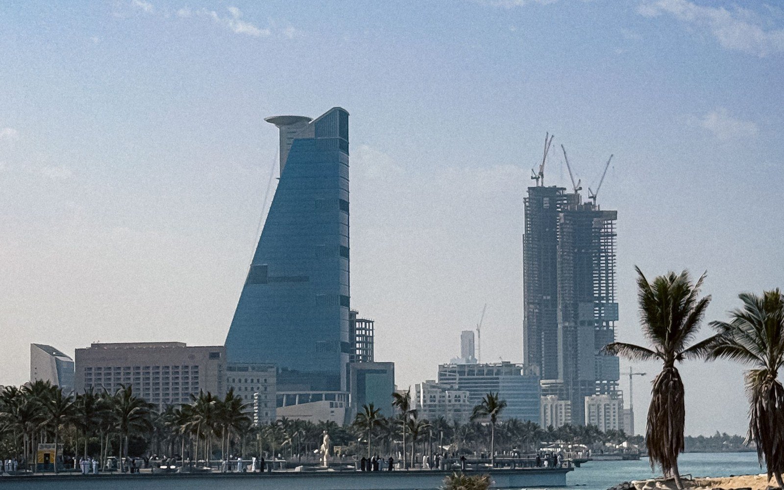 The Jeddah waterfront seen from the sea, with a tapering glass tower, a second tower under construction with four tower cranes on it, low buildings and palm trees along a rock breakwater