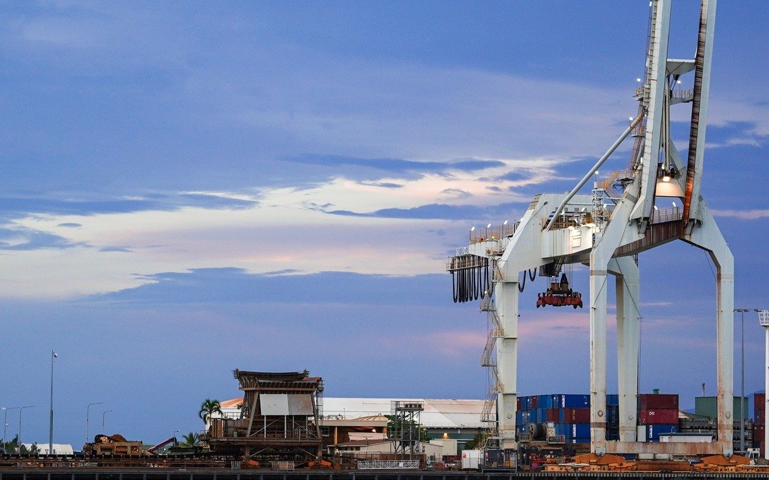 A ship-to-shore container crane on a quay at dusk, its spreader lowered between the legs, with stacked containers and storage tanks behind and open water in the foreground, location not identified
