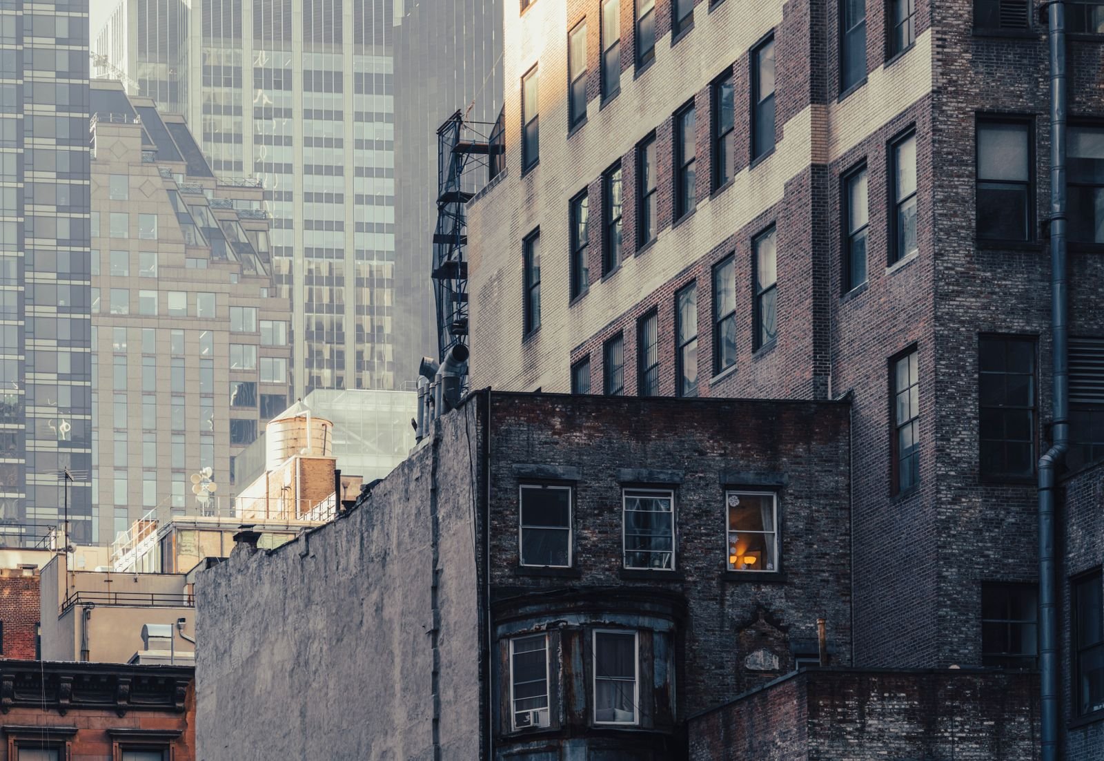 Brick and stone buildings of different eras packed together in New York at dusk, with taller towers behind them in the haze