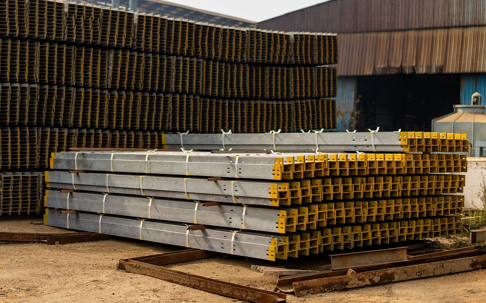 Bundles of galvanised steel sections banded and stacked in an open yard, bright zinc coated members with punched holes alongside yellow primed sections, photographed square to the ends