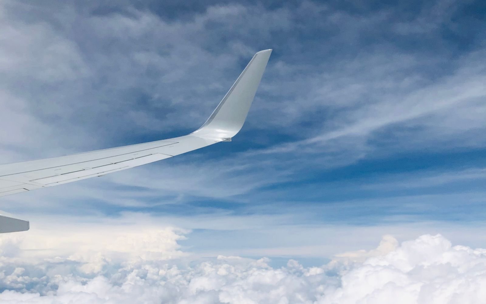 The white wing and upswept winglet of an aircraft in flight above a broken layer of cloud under a blue sky