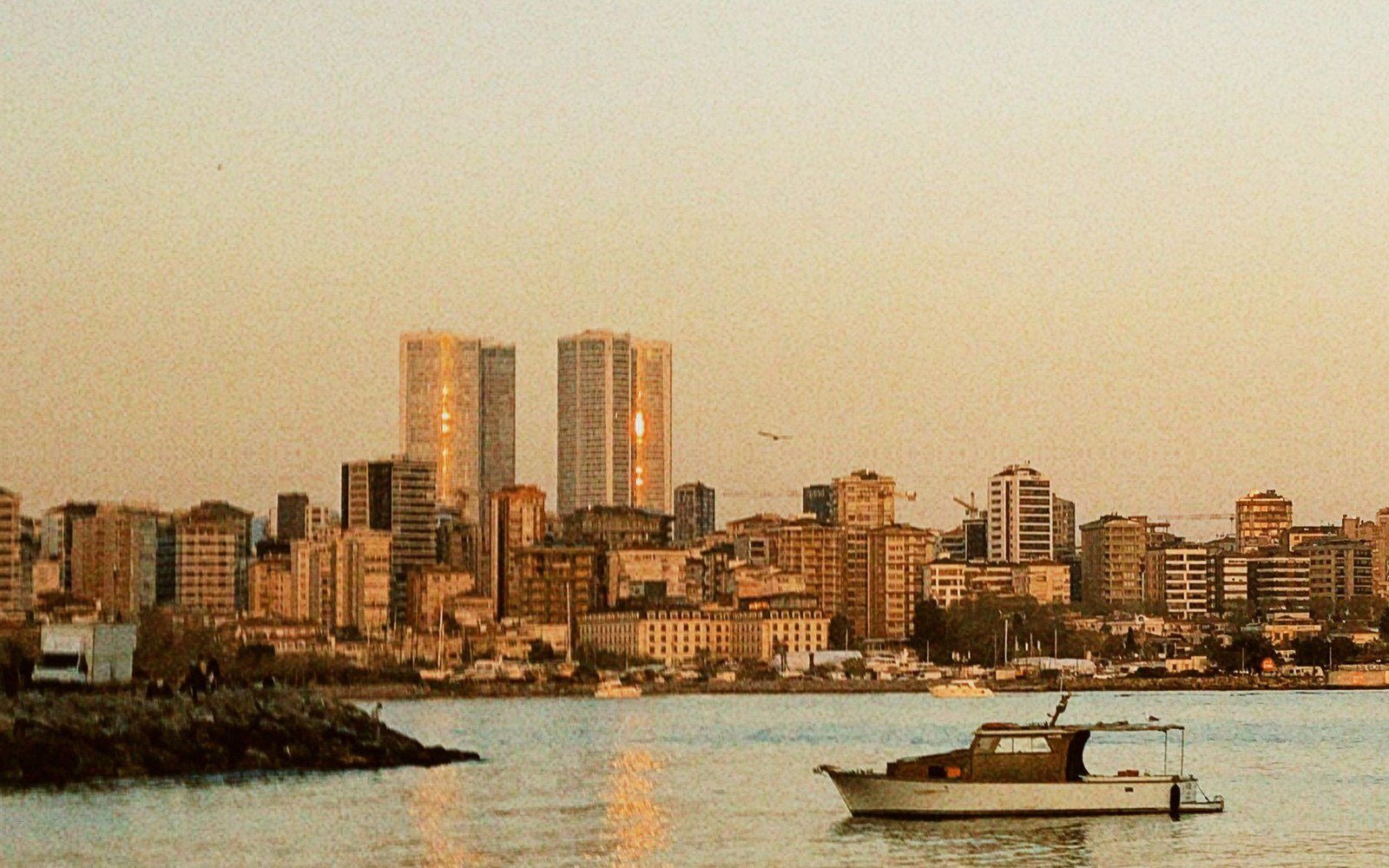 The Beirut waterfront at sunset seen across the water, with two towers standing above a dense line of apartment blocks and a small boat moored in the foreground