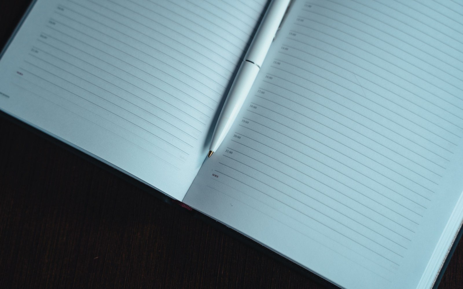 An open ruled notebook with a slim white pen resting in the gutter, lying on a dark wooden desk