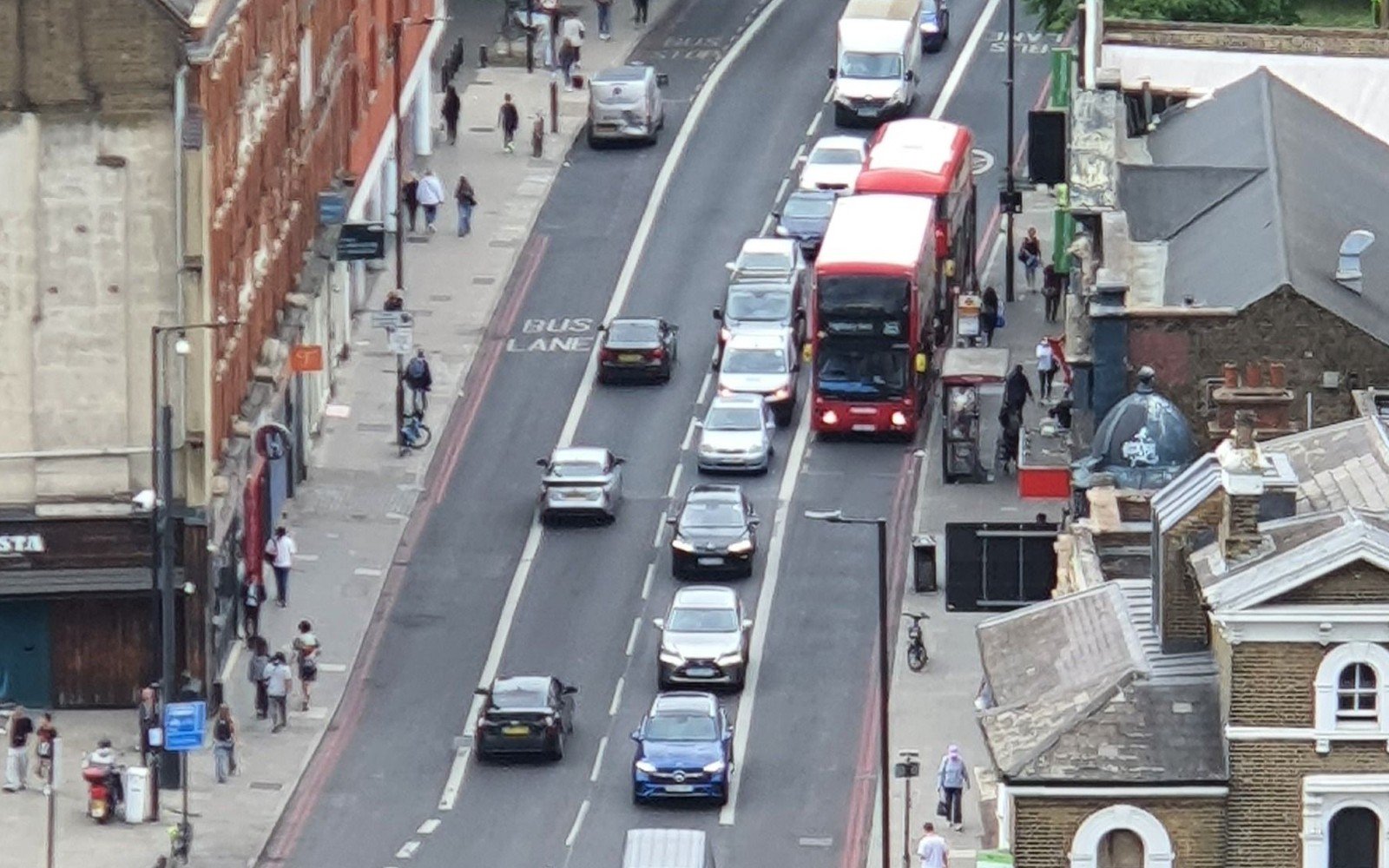 An aerial view down a busy London street, cars and a red double decker bus queueing in two lanes with a marked bus lane alongside and terraced buildings on both sides