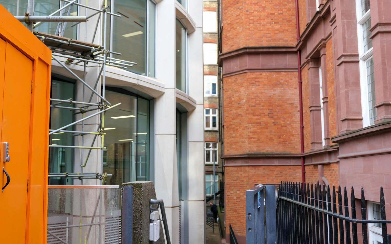 A new building under construction behind site hoarding and scaffolding, standing immediately alongside an older red brick building