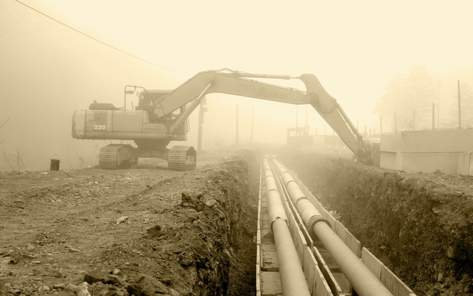 An excavator standing beside an open trench with two large diameter pipes laid along its base in hazy light