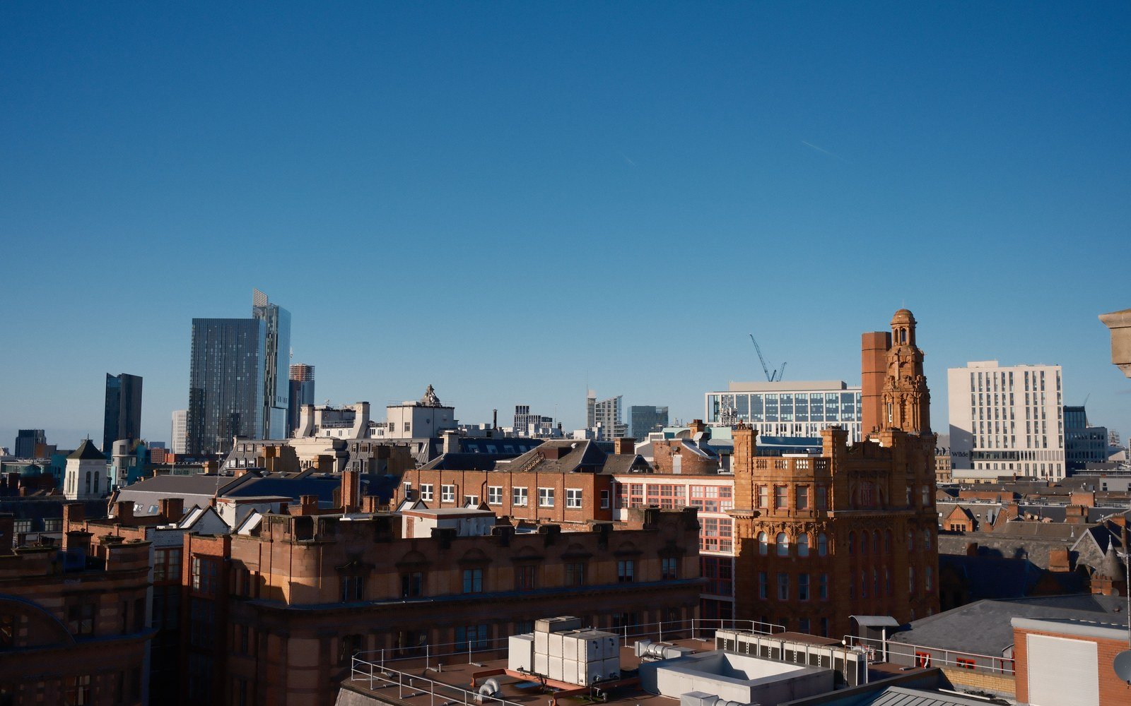 Manchester rooftops in low sun, with Victorian brick facades in front of glass towers and a tower crane on the skyline