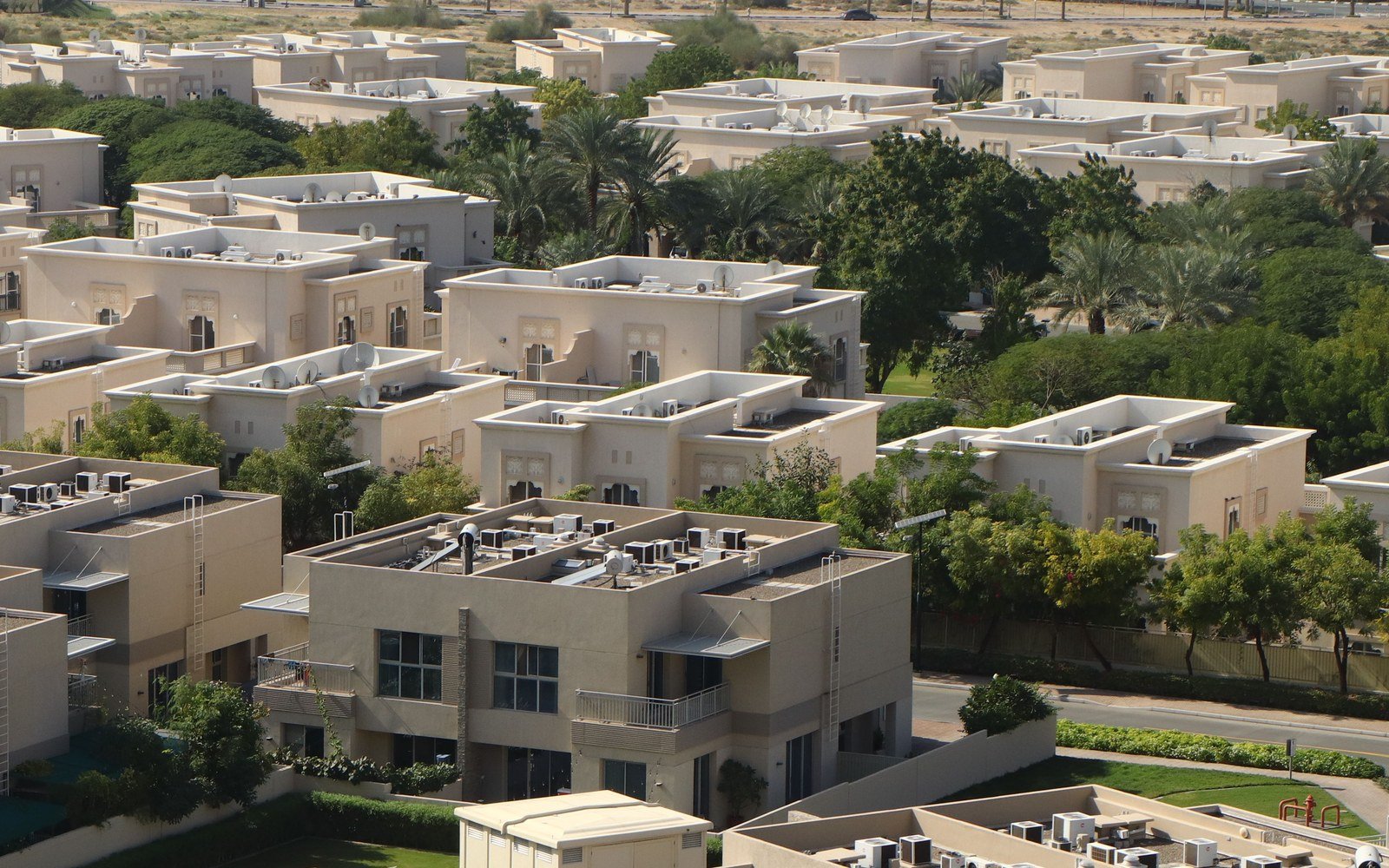 Daylight aerial view over a low rise residential community of flat roofed beige villas and town houses, with rooftop plant, mature trees and planted verges between the streets
