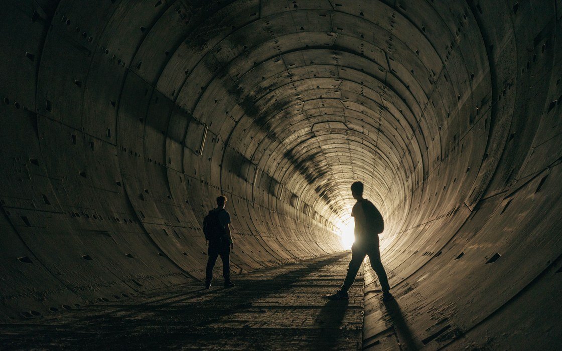 The inside of a bored tunnel lined with precast concrete segment rings, with two figures in silhouette against daylight at the far end