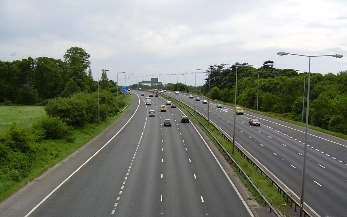 A three lane motorway curving through a wooded cutting, with lighting columns, safety barriers and lane markings