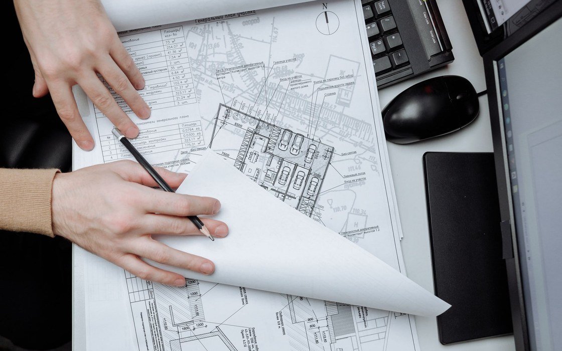 Two hands unrolling a technical floor plan across a desk beside a keyboard, scale rule and pens